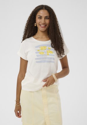 Femme souriante aux longs cheveux bouclés portant un t-shirt blanc imprimé d'une fleur jaune et une jupe jaune clair, accessoirisée avec des bijoux en or et un bracelet.