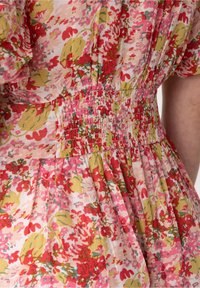 Květinové šaty s růžovým, zeleným a červeným vzorem a nařaseným pasem. Materiál je lehký a mírně průhledný s plisovanými detaily.