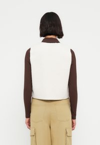 Veste en peau lainée blanche à la coupe courte, portée par-dessus un top marron à manches longues côtelées. La tenue est complétée par un pantalon cargo beige.