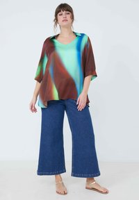 Flowy, silk top with a v-neck, featuring a green, brown, and blue abstract pattern. Paired with wide-leg blue denim jeans and sandals.
