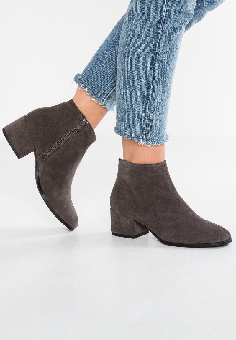 Bottes chevilles en daim gris foncé avec talons blocs et fermetures éclair latérales, portées avec des jeans bleu clair courts et effilochés sur un sol blanc.