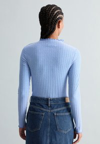 Personne aux cheveux tressés portant un haut à manches longues côtelé bleu clair et une jupe en jean bleu foncé taille haute, vue de dos.