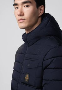 Giovane uomo che indossa un giubbotto trapuntato blu navy con colletto alto e un logo a patch sulla tasca del petto.