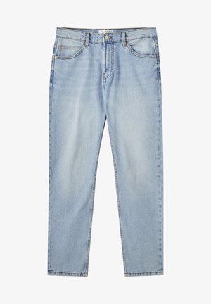 Person iført lyseblå jeans, grå oversized T-shirt med brystlomme, hvide og grå sneakers, der viser en farverig tatovering på venstre underarm.