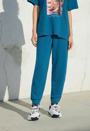 Personne portant un pantalon de survêtement bleu sarcelle et un t-shirt oversize assorti avec un motif de baskets, debout sur un sol en béton contre un mur blanc.