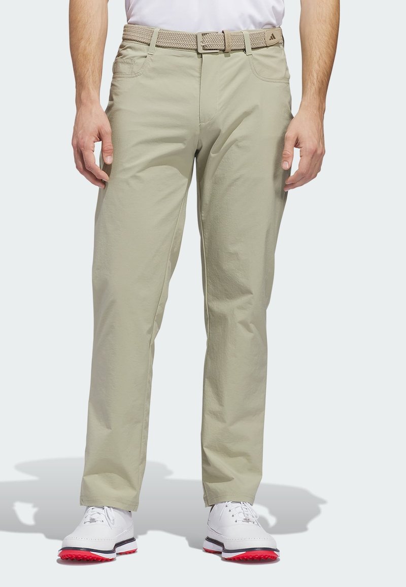 Άνδρας με μπεζ casual παντελόνι και υφαντή ζώνη, φορώντας λευκά παπούτσια γκολφ με κόκκινες σόλες, στέκεται μπροστά σε απλό φόντο.