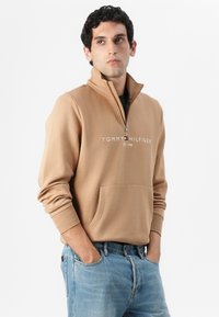Beige mikina na zips s predným vreckom, s logom "TOMMY HILFIGER EST. 1985" v bielej farbe, nosená cez čiernu košeľu, skombinovaná s modrými džínsami.