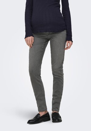 Person iført tætsiddende grå jeans, sorte loafers og en mørkeblå ribbet top med lange ærmer, stående med afslappede hænder.