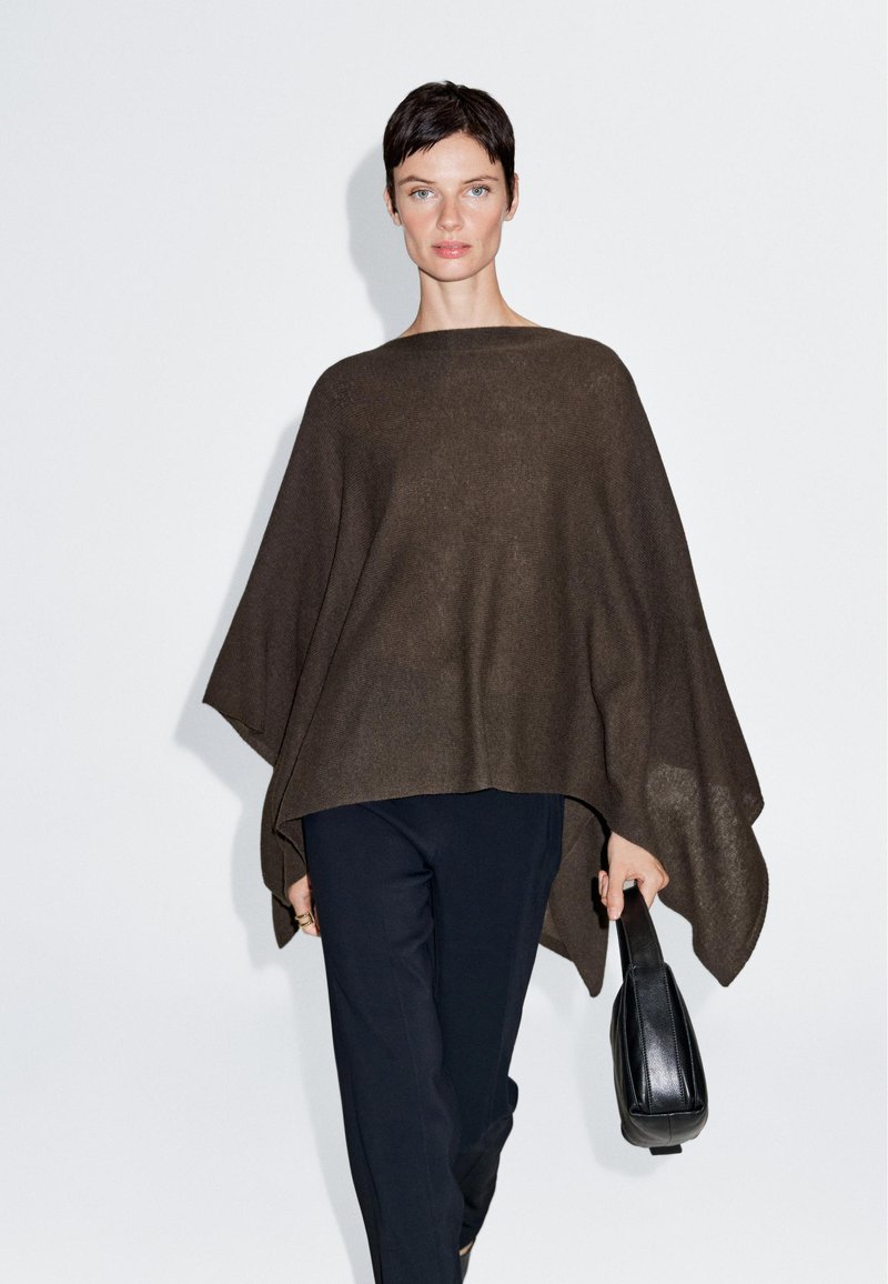 Dunkelbrauner Poncho aus weichem, strukturiertem Stoff, mit einer breiten, fließenden Form. Kombiniert mit dunklen Hosen und einer kleinen schwarzen Handtasche.