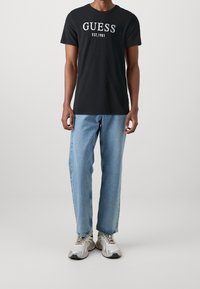 Sort bomulds T-shirt med hvid "GUESS EST.1981" grafik. Parret med lyseblå, løstsiddende denim jeans og sølvfarvede sneakers.