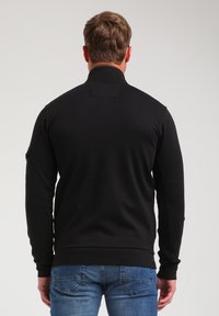Felpa nera con zip, colletto alto, polsini a coste e tasca laterale. Texture liscia e design aderente, mostrata da dietro.