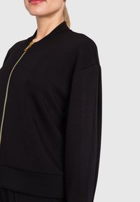 Giacca bomber nera con zip, dal tessuto morbido, caratterizzata da un colletto alto e una cerniera in tono oro. Design elegante e semplice, vestibilità su misura.