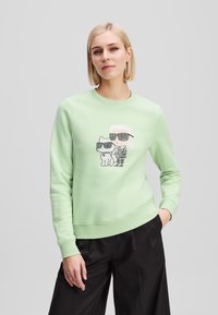 Felpa verde chiaro con maniche lunghe, caratterizzata da una grafica di un personaggio e un gatto con occhiali da sole, con polsini e orlo a costine.