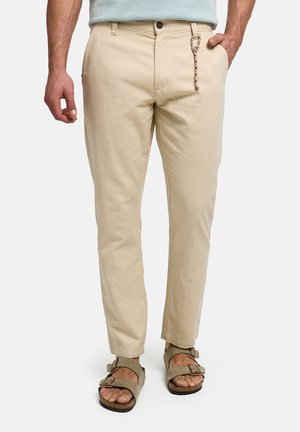 Mand iført beige chinos med en flettet nøglering, lyseblå skjorte og brune sandaler med to stropper, stående mod en hvid baggrund.