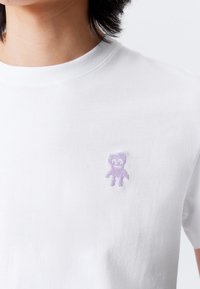 White cotton t-shirt with a round neckline and short sleeves. Features a small lavender embroidered bear design on the left chest.