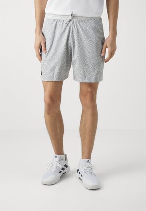 Helle graue Shorts mit einem abstrakten, wellenförmigen Muster. Sie haben einen elastischen Bund und Seitentaschen. Kombiniert mit weißen Sportschuhen.