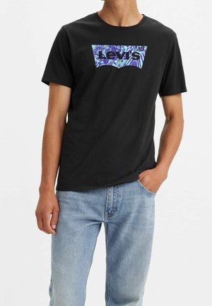 Mann trägt schwarzes Levi’s-T-Shirt mit blauem Blumenlogo und hellblauen Jeans, rechte Hand in der Tasche, vor weißem Hintergrund.