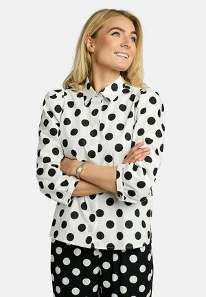 Bijela košulja s gumbima i velikim crnim točkicama, s prstenastim manžetama i klasičnim ovratnikom. Model prekriženih ruku prikazuje kroj košulje.