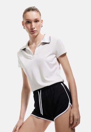 Valkoinen polo-paita mustalla kauluskivellä, yhdistettynä mustiin urheilushortseihin, joissa on valkoinen reuna ja nauhakuulunta. Sileä kangas.