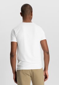 T-shirt bianca in cotone con collo rotondo, maniche corte e design semplice. Indossata con pantaloni beige, mostrando una silhouette aderente da dietro.