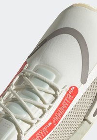 Vit idrottssko med grå böjd design, vita snören, mesh-sida och röd etikett med texten "NMD:ATA FLOW" på sidopanelen.