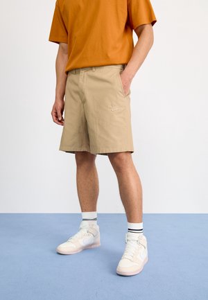Mężczyzna w beżowych spodenkach Nike, białych wysokich butach sportowych z białymi skarpetkami w czarne paski oraz pomarańczowej koszulce z krótkim rękawem stoi na niebieskiej podłodze.