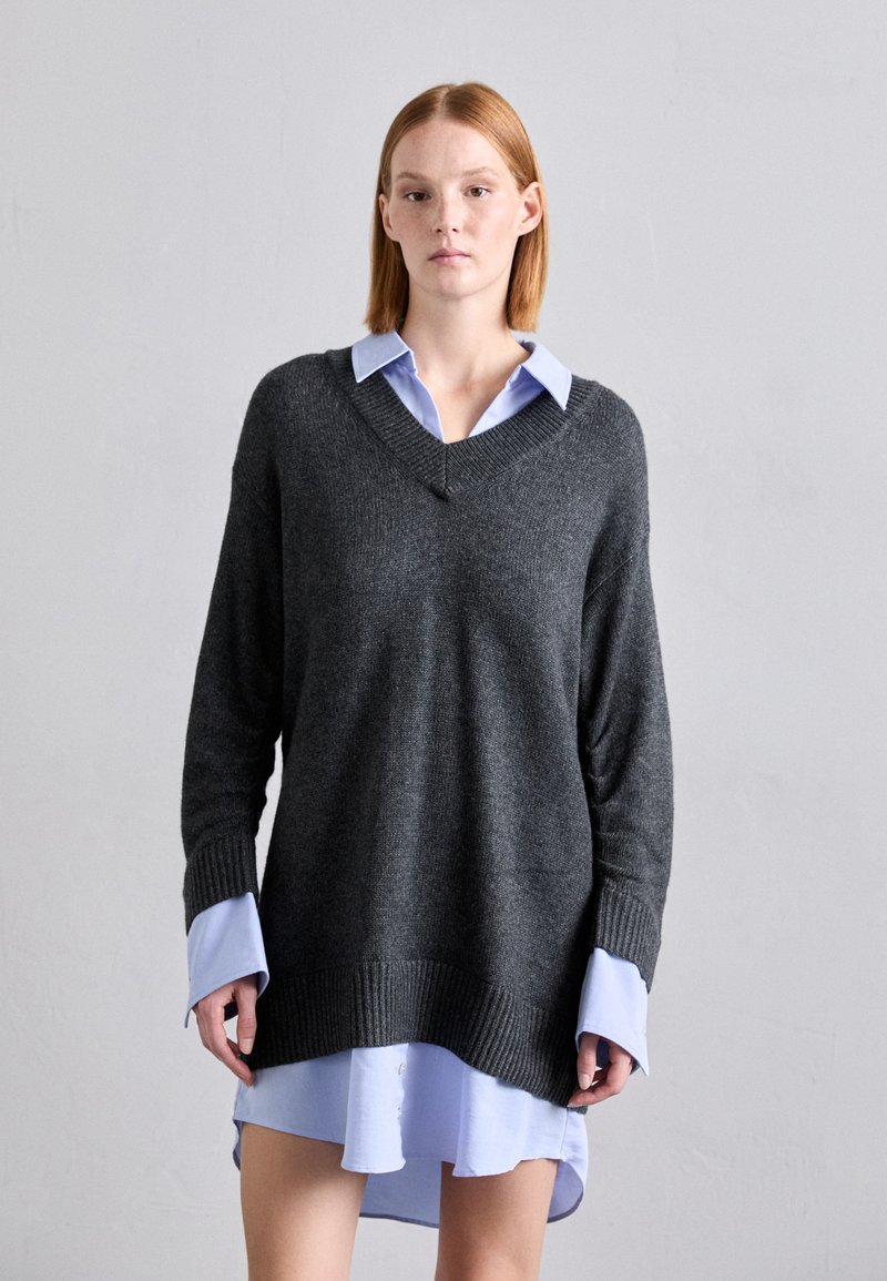 Pulover gri din tricot, cu un design în formă de V și mâneci lungi. Purtați-l peste o cămașă albastră deschisă, cu guler, care are detalii cu nasturi și un tiv curbat.