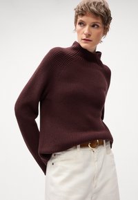 Maglione a coste bordeaux con colletto alto, abbinato a pantaloni bianchi e una cintura marrone con fibbia dorata.