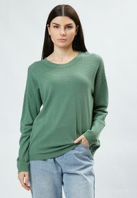 Groene pullovertrui met een textuurgebreid patroon op het bovenste gedeelte, geribbelde mouwen en een relaxte pasvorm, gedragen met lichtblauwe jeans.