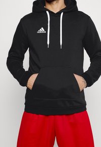 Sweat à capuche noir en tissu doux, avec une poche avant et des cordons blancs. Un petit logo Adidas blanc est présent sur le haut à gauche de la poitrine.