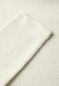 Tejido blanco con una textura suave, que presenta un puño doblado y un dobladillo limpio. El fondo muestra un patrón sutil de canalé.