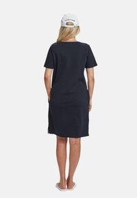 Rochie scurtă, de culoare bleu marin, cu mâneci scurte și o croială lejeră, dotată cu buzunare laterale. Include o șapcă albă cu detalii logo. Materialul pare moale și neted.