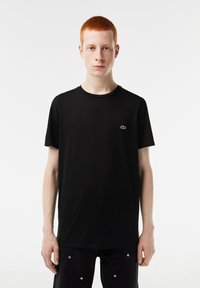 Camiseta de algodón negra con cuello redondo, mangas cortas y un pequeño logo de cocodrilo verde en el lado izquierdo del pecho. El modelo está en una pose neutral.