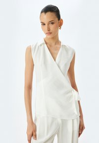 Witte mouwloze blouse met een kraaghalslijn, wikkeldesign, een zijstrikdetail en zachte, gestructureerde stof. Gepaard met bijpassende lichte broek.