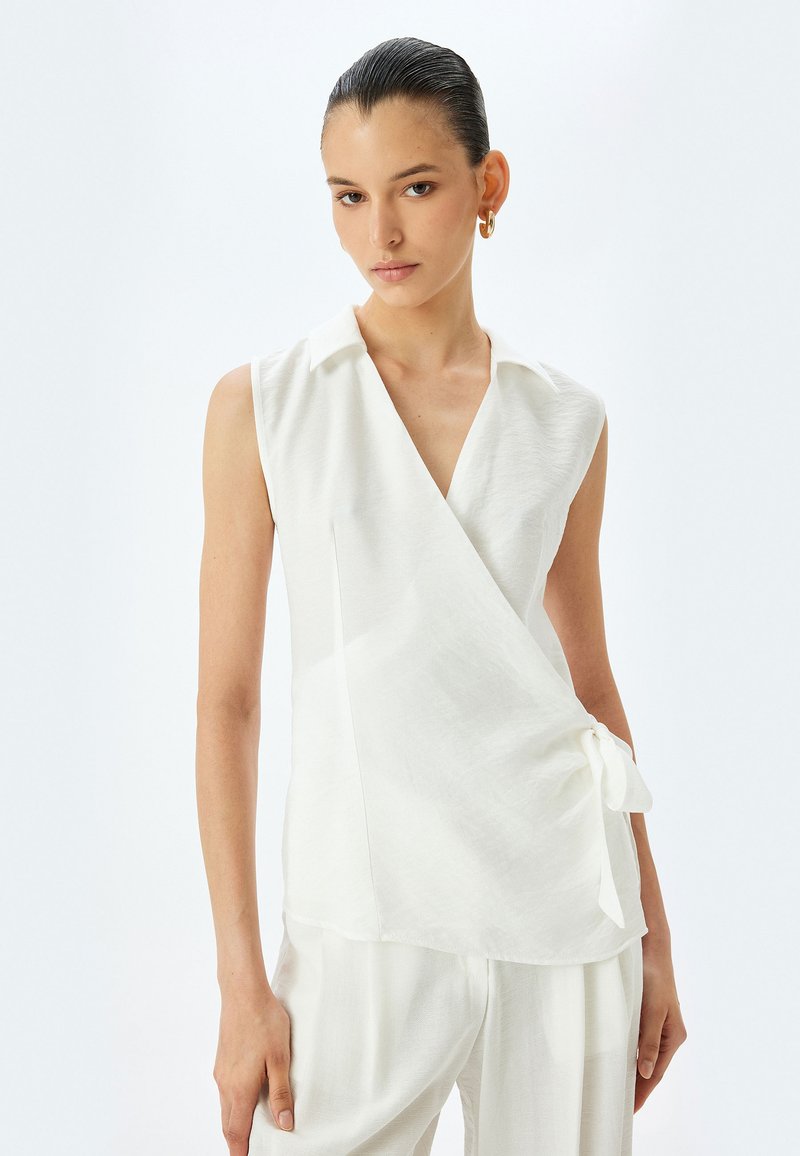 Witte mouwloze blouse met een kraaghalslijn, wikkeldesign, een zijstrikdetail en zachte, gestructureerde stof. Gepaard met bijpassende lichte broek.