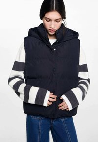 Ullväst i marinblått med avtagbar huva, med knappar och quiltad design, bärs över en vit randig tröja.