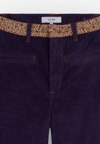 Pantalon en velours côtelé violet foncé avec des coutures orange et une ceinture beige à imprimé léopard passée dans les passants.
