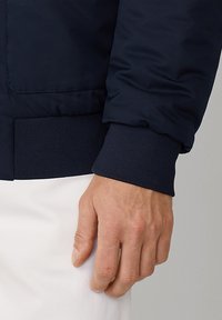 Morska niebieska kurtka bomberka z mankietem o prążkowanej fakturze, z gładkim wykończeniem tkaniny. Ręka spoczywa na białym elemencie odzieży poniżej.