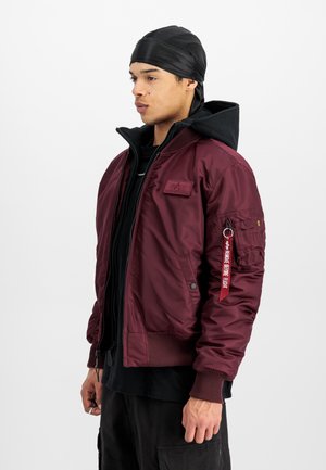 Maroon bomber jacket with a black hood, ribbed cuffs, and a zip pocket on the sleeve. Features a tag with text and a smooth finish.