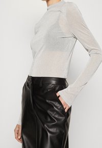 Kvinna iklädd en långärmad topp i silverglitter instoppad i höga svarta läderbyxor med en hand i fickan.