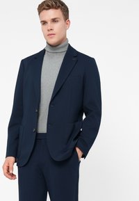 Mladý muž v tmavomodrom saku a zodpovedajúcich nohaviciach s svetlošedým rolákom, stojí s jednou rukou v vrecku.