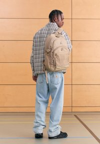 Mochila bege com vários compartimentos com fecho, com o logótipo da Jordan. Usada sobre uma camisa xadrez clara e jeans azuis largos.