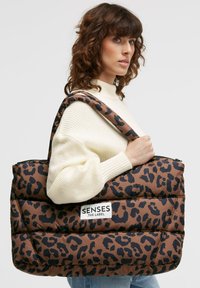 Gepolsterte braune Tragetasche mit Leopardenmuster und großem weißen Logopatch. Verfügt über einen breiten Schultergurt und einen Reißverschluss oben. Glatte Oberfläche.