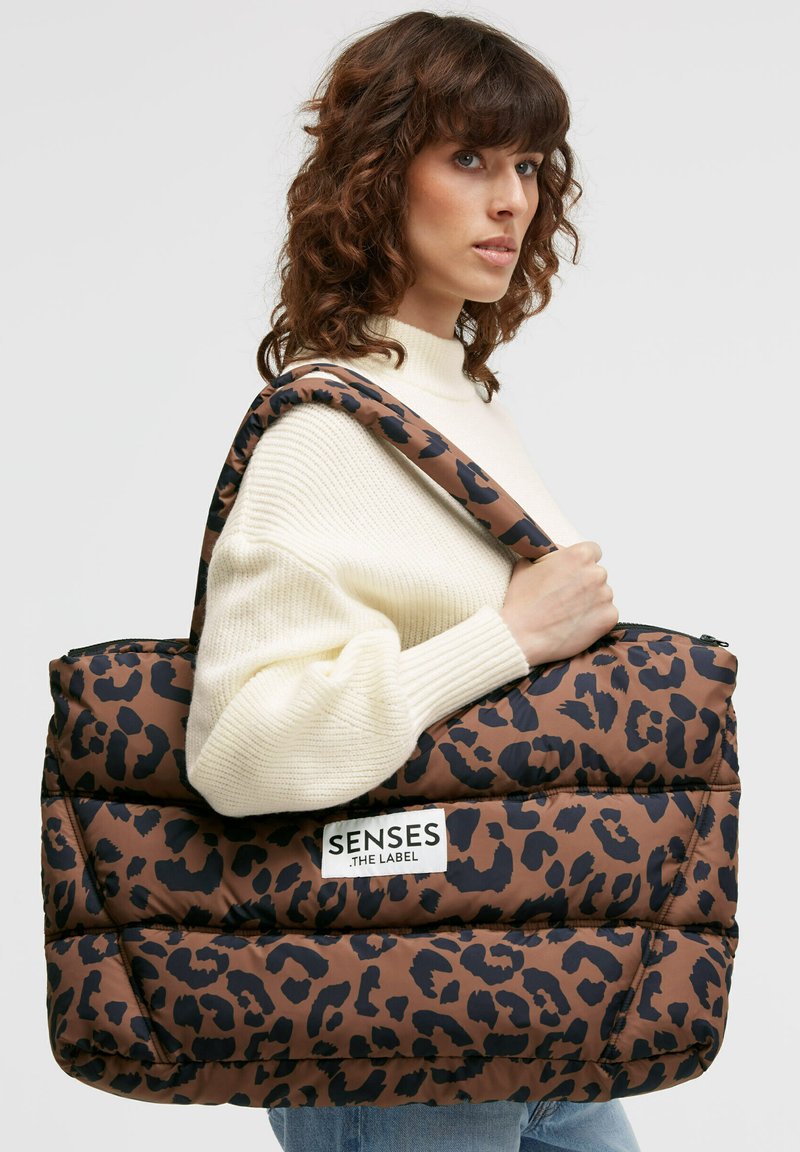 Gepolsterte braune Tragetasche mit Leopardenmuster und großem weißen Logopatch. Verfügt über einen breiten Schultergurt und einen Reißverschluss oben. Glatte Oberfläche.