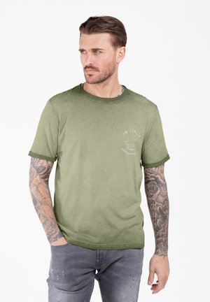 Homme avec des bras tatoués portant un t-shirt vert avec l'inscription "In Italia" et un jean gris déchiré, se tenant avec une main dans la poche, regardant sur le côté.