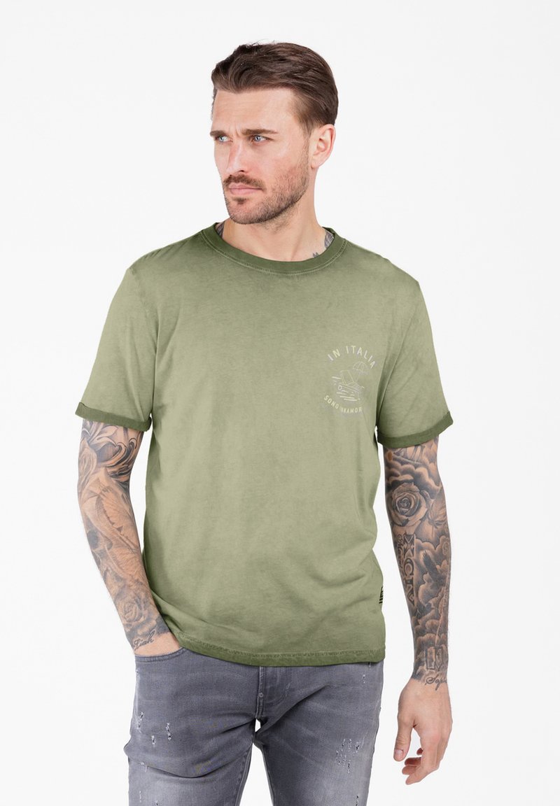 Hombre con brazos tatuados que lleva una camiseta verde con la impresión "En Italia" y unos jeans gris desgastados, de pie con una mano en el bolsillo, mirando de lado.