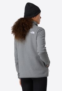 Persona con capelli ricci che indossa un berretto nero, una giacca in fleece grigia con zip e il logo The North Face, e pantaloni neri, di spalle con le mani nelle tasche.
