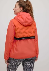 Červenooranžová péřová bunda s kapucí, která má kontrastní černý pruh v pase. Textura zahrnuje hladké a prošívané části.