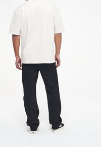 Krémový oversized tričko s krátkým rukávem s nápisem "PEGADOR" na zádech, v kombinaci s černými kalhotami rovného střihu a černými teniskami.