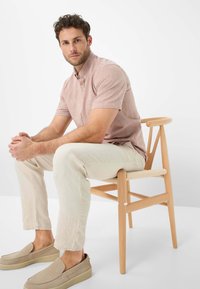 Camicia rosa chiaro a maniche corte con bottoni, pantaloni in lino crema e scarpe beige senza lacci, seduto su una sedia di legno con seduta intrecciata.